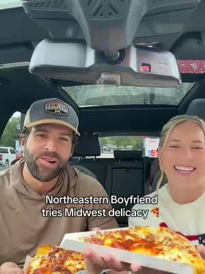 A man and a woman are sitting in a car, holding and looking at a pizza. The man is wearing a cap and the woman smiles widely, with pizza in the foreground.