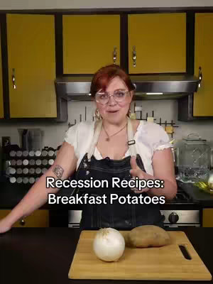 A medium shot shows a woman in a kitchen setting. She is standing in front of the countertop and a yellow cabinet behind her. She is wearing glasses and an apron and has the kitchen utensils on the countertop. There is a whole onion and a potato.