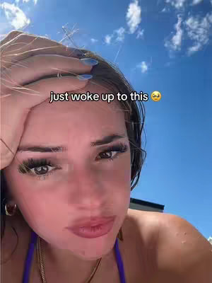 A selfie-style video appears with a young woman's face filling much of the frame. Her hand is resting on her forehead, and the background is a bright blue sky with wispy clouds. The woman's expression suggests a mix of surprise and affection.