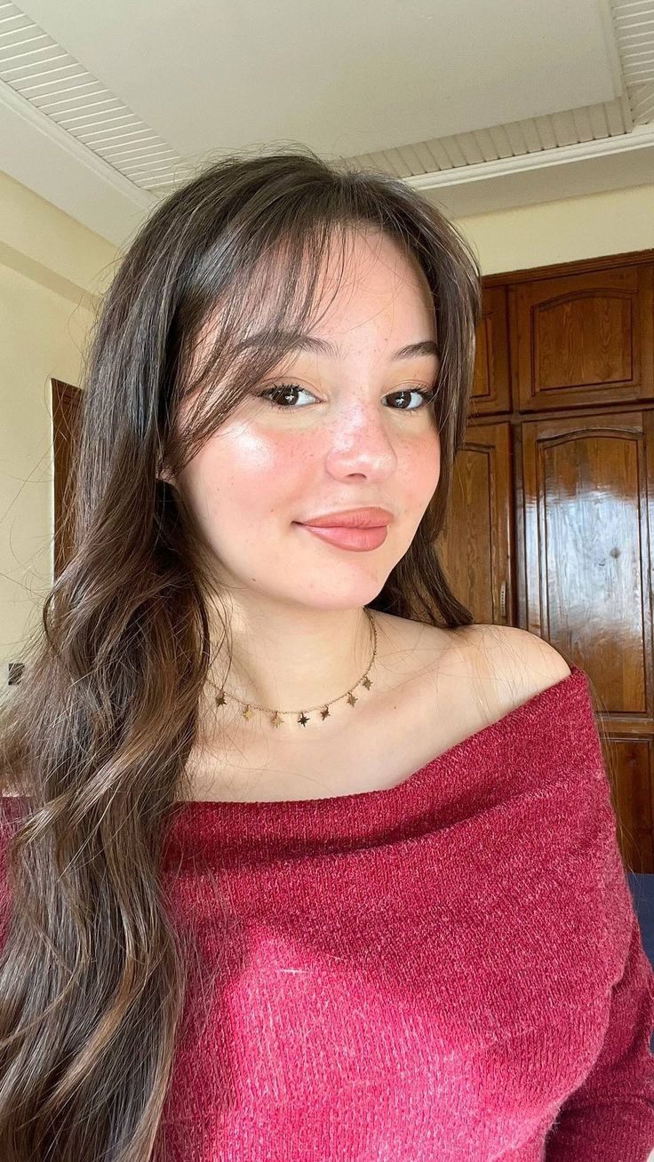 A portrait of a young woman with long brown hair and soft, freckled skin. She is wearing a red off-the-shoulder sweater and a delicate star-shaped necklace. Her expression is a subtle smile. The background includes wooden cabinets and a wall, with natural light illuminating the scene.