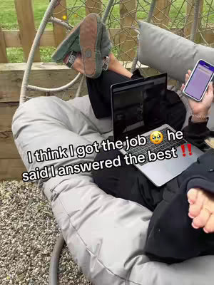 A person is relaxing outside while interacting with multiple devices. They are in a hanging chair, and a laptop is balanced on their lap. They hold a mobile phone in the right hand. The person is wearing a black attire and putting their feet, with slippers on, up in the air.