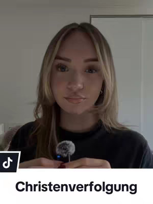 A woman with light blonde hair is centered in the frame, looking towards the camera, which is positioned close-up. She holds a microphone in her hand. The background is a neutral, light-colored room.