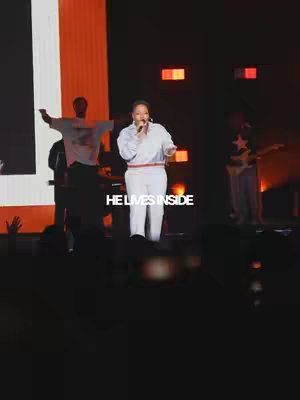 A live performance video featuring a woman in white singing with a microphone. The background includes a stage setting with other performers, lighting, and an audience, and the text overlay 'HE LIVES INSIDE'.