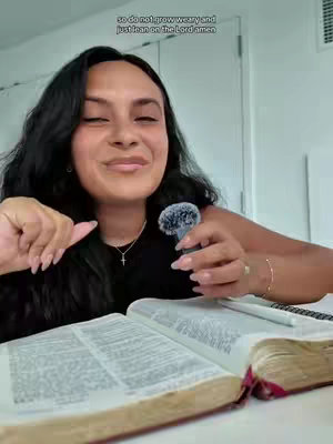 A woman with long dark hair sits at a table, smiling and gesturing towards a microphone. She's holding a Bible open in front of her with a pen, suggesting a message about faith and reflection. The background is a simple white wall.