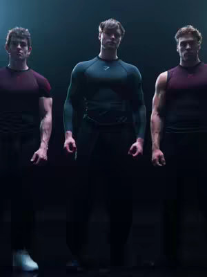 Three muscular men stand in a dimly lit setting, posing with athletic wear. The central figure is highlighted with a brighter light.