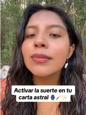 A medium shot of a woman with dark wavy hair, looking at the camera. The background is blurred green foliage. She has light skin, and is wearing a reddish-orange top. A text overlay at the bottom states: "Activar la suerte en tu carta astral" with an eye and a pencil symbol.