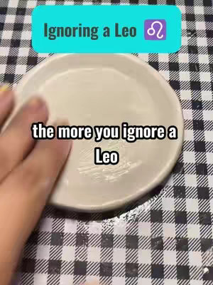 The image features a close-up perspective of a white, round plate with a hand lightly touching the surface. A tablecloth with a black and white checkered pattern provides the background. Two overlaid text boxes are present: one at the top with the text 'Ignoring a Leo' along with the Leo zodiac symbol, and another at the center containing the text 'the more you ignore a Leo' in a sans-serif font