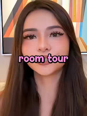 A woman with long, dark brown hair and makeup is centered in the frame. The main subject appears to be in the room in which the video is being filmed. The background is a bit blurred, with a decorative element peeking out on the right.