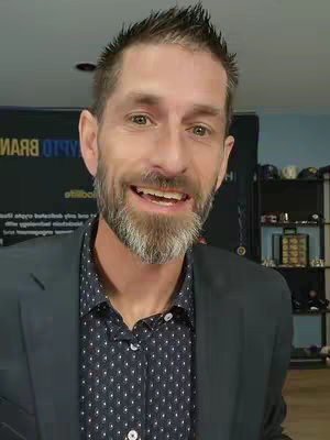 A medium shot of a man facing the camera. He has short, dark hair and a beard with some graying. He is wearing a dark patterned shirt under a dark blazer. Behind him is a blurred background with a shelf displaying items and what appears to be a promotional backdrop with text.