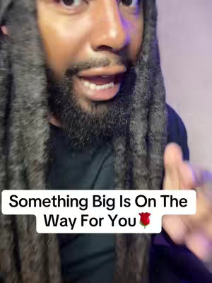 A close-up shot of a Black man with long dreadlocks, a beard, and a serious expression. He is wearing a black top, and a white text overlay at the bottom states, "Something Big Is On The Way For You" followed by a red rose emoji. The background is blurred, and the lighting is soft.