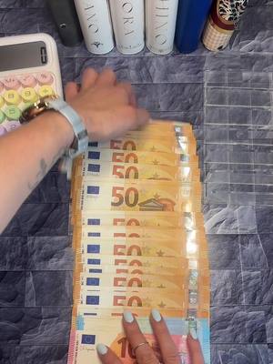 The image shows a person's hands arranging a stack of Euro banknotes, primarily 50 Euro notes, on a patterned surface. Other items, such as a calculator, makeup, and stationery, are visible in the background.