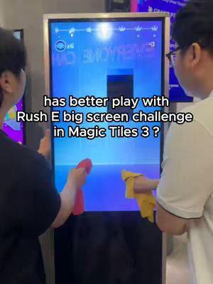 The image shows two people, viewed from behind, flanking a large screen displaying a music game interface. Text overlays describe the game challenge. They are holding red and yellow cloths, potentially for cleaning the screen.