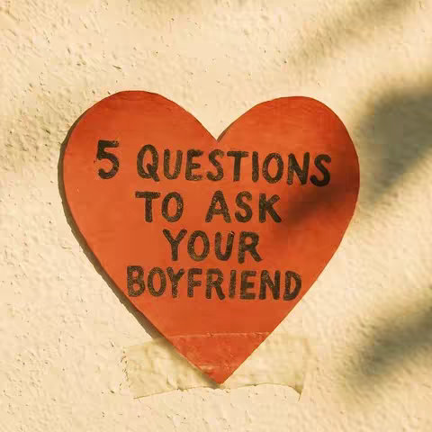 A close-up shot of a red, heart-shaped sign with the words '5 QUESTIONS TO ASK YOUR BOYFRIEND' in black letters. The sign is attached with tape to a lightly textured, off-white wall. The lighting is soft, with slight shadows.