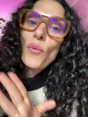A close-up shot of a woman's face with curly dark hair, wearing large, square-framed glasses. Her lips are slightly parted, and she has a neutral expression.