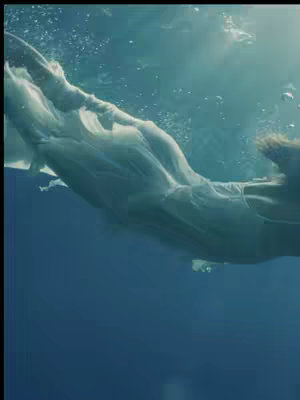 The image shows a person submerged in water, with their body and clothing partially visible. The scene is underwater, with bubbles and light rays adding to the ethereal feel. The person's clothing billows softly, appearing to be white or light-colored, which contrasts with the deep blue water.
