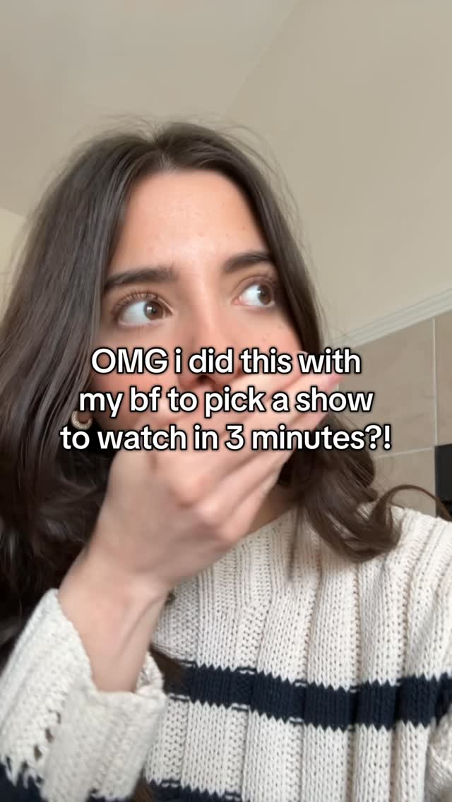 The image is a selfie of a person covering their mouth with their hand. The person is a woman with long, dark hair and fair skin. She is wearing a cream-colored knit sweater with navy stripes. Text overlayed on the image reads: Omg i did this with my bf to pick a show to watch in 3 minutes?! The background is neutral, with a hint of a white wall. The composition is centered on the woman's face, creating a sense of intimacy and direct engagement.