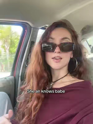 A woman with long red hair, wearing sunglasses and a maroon top, sits in a car. The background has trees and sunlight visible through the window. Text overlayed on the image.