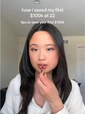 A selfie-style video cover. A young Asian woman with long dark hair is applying lipstick. Above her, text overlays state 'how i saved my first $100k at 22' and 'tips to save your first $100k'