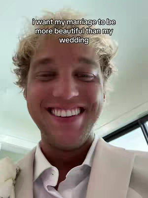 A close-up shot features a smiling man with blonde hair wearing a white suit and holding a white flower. Text overlay declares, "i want my marriage to be more beautiful than my wedding". The composition focuses on the man's face with a background suggesting an indoor setting.