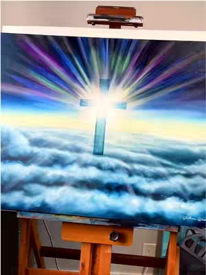 An oil painting on a wooden easel. The painting depicts a bright, glowing cross against a background of fluffy clouds and radiating beams of light in shades of blue, purple, and white. The background is a gradient of blues and yellow. The overall composition is vertical and centered.