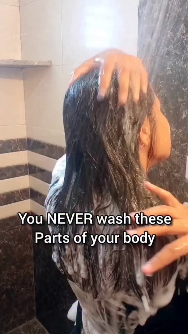A woman is in a shower, and the upper portion of her body is visible beneath the spray of the shower. She is washing her hair with both hands, with soap visible. Text in the bottom says "You NEVER wash these Parts of your body".