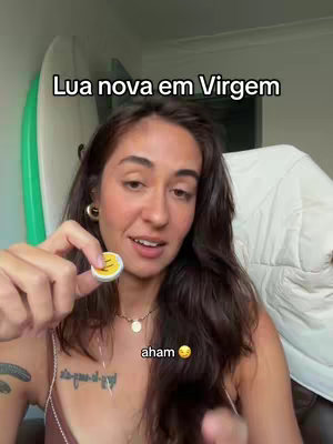 A woman with long, dark hair smiles and holds up a small circular item with a yellow smiley face on it. She is wearing a necklace and has a tattoo on her arm. The background is a light-colored wall with a white object in the corner, and a surfboard. Text overlays at the top and bottom of the image. The overall composition is well-lit and focuses on the subject.