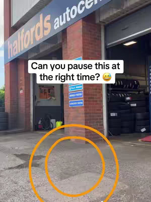 The image shows the exterior of a Halfords Autocentre. The main focus is on a paved area in front of the service bay. A large orange circle is drawn to highlight a specific area, likely related to a challenge described in the caption. There are also some tires.