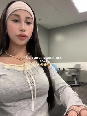 A selfie-style video cover featuring a young woman with dark hair, wearing a headband and a light-colored top. The woman is smiling slightly and looking towards the camera. Text overlayed on the picture reads "POV: YOU'RE IN OB LECTURE DAY 1," along with baby, chick, and teddy bear emojis.