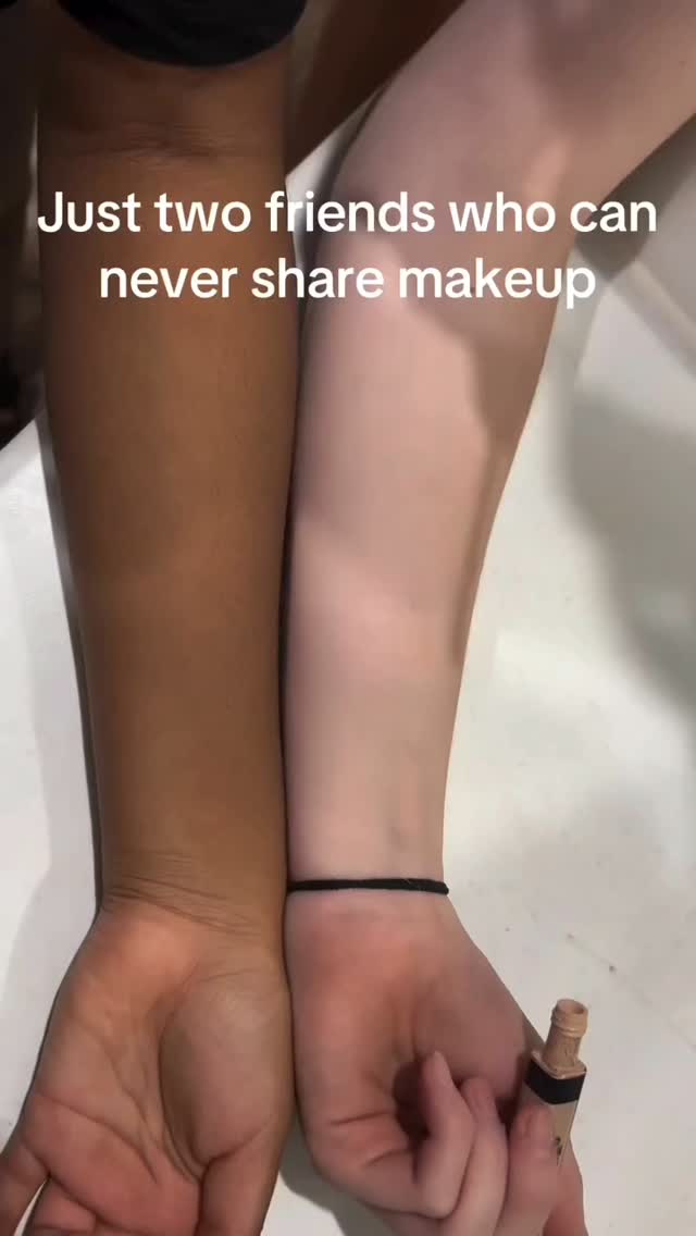 The image shows two arms, one with a dark skin tone and the other with a light skin tone, held side by side against a white background. The hands are open. The text overlay reads Just two friends who can never share makeup. A tube of concealer is in one of the hands.