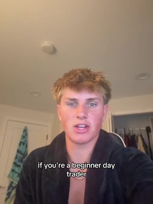 A selfie-style video cover image featuring a young man with short, wavy blonde hair. He's wearing a dark, draped robe or garment and a delicate gold chain. The background is a softly lit room with off-white walls and a recessed ceiling light. Text overlayed on the image reads, "if you're a beginner day trader".