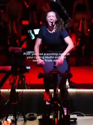 A woman is shown on a cycling bike inside a cycling studio. She is wearing a dark top and cycling pants. She is singing with her mouth open. The scene is lit with red lights.Text overlay in white font says, 'POV: you had a worship night at your cycling studio and this was the finale song.'