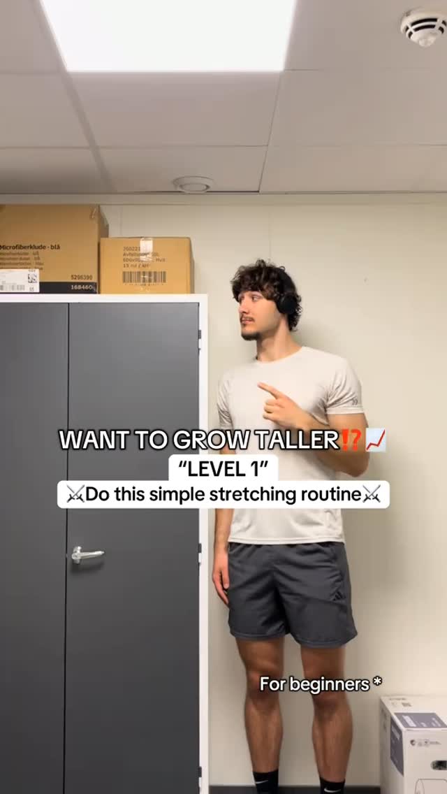 A man in a light colored t-shirt and gray shorts stands in a room with a dark gray door and various boxes. Text overlays on the image ask if the viewer wants to grow taller and offer a level 1 stretching routine marked as suitable for beginners.