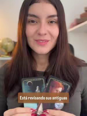 A woman with long brown hair is holding tarot cards. A text overlay states 'Está revisando sus antiguas'. The background shows a globe and parts of a shelf.