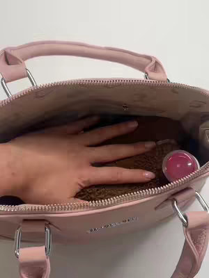 A light pink purse, partially open, with a hand inside. The purse appears to be leather or a similar material. The background is white. There is a pink item and some items in the purse.