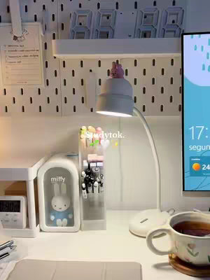 A cozy desk setup with a white desk and shelves against a white wall. There's a computer on the right, a lamp with a small figure on top of the bulb, pens and markers in a clear holder, and a mug of tea. The overall aesthetic is clean and inviting.