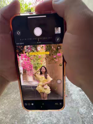 A person holding a phone in their hands, capturing a photo of a woman in a yellow dress. The phone's screen displays the camera interface, showing the woman in the center, posing against a backdrop of pink flowers and a pathway. The scene is framed with the hands and the phone's orange case visible. The background is blurred to emphasize the main subject of the photo, located outdoor.