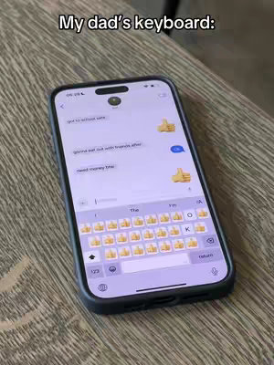 A phone displaying a text message conversation is resting on a wooden surface. The phone's screen shows a chat with messages and thumbs-up emojis, which seem to be a humorous take on a father's texting style. The phone is in a dark case, and the background shows wooden grain.