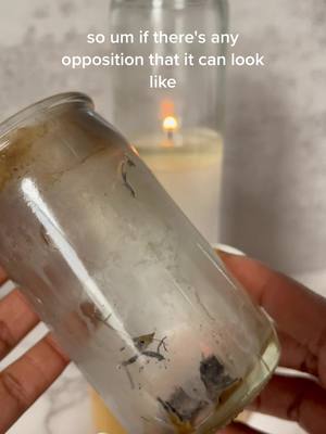 The image shows a close-up of a clear glass jar being held at eye level. Inside the jar are dark, charred remnants. Behind the jar, a second candle is burning. The background appears to be an off-white wall.