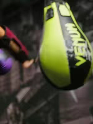 The image shows a person's arm in the act of kicking a neon green and black punching bag. The bag has the word 'VENUM' in yellow. The background is blurry and seems to be an industrial style wall.