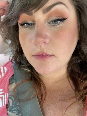 An up-close selfie of a woman with fair skin, auburn hair, and light-blue eyes. She is wearing makeup. The background is slightly out of focus. She is wearing a pink shirt. There is a seatbelt in the image.