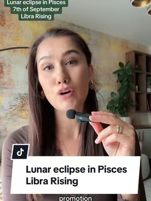 A woman with brown hair is facing the camera, holding a microphone. There is text overlayed on top of the image that states: Lunar eclipse in Pisces, 7th of September, Libra Rising. The video is filmed indoors, with a neutral background.