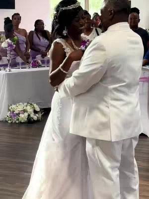 A wedding scene featuring a bride and groom dancing. The bride in a white gown and tiara looks lovingly up at the groom wearing a white suit. Guests are visible in the background at tables.