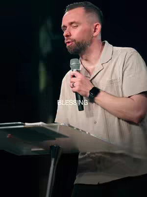 A man with short hair and a short beard is on a stage with a microphone in his hand. He is giving a speech. There is text at the bottom of the screen that says 'BLESSING'. The shot is at a medium proximity, and the background is dark.