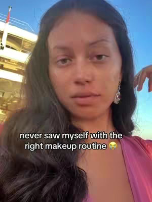 A woman with long, dark hair looks directly at the camera. She's wearing a pink top. Behind her, a ship is faintly visible, suggesting an outdoor setting. Text overlayed on the image reads, "never saw myself with the right makeup routine". The overall impression is one of self-awareness within a beauty context.