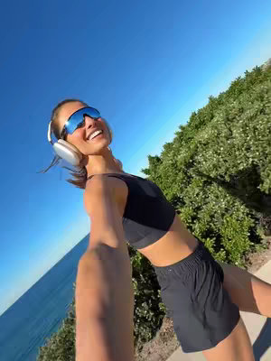 A selfie-style shot taken from slightly below the creator. The creator, a woman with dark hair, is smiling broadly and angled slightly to the right. She's wearing blue mirrored sunglasses and silver headphones. She's dressed in a black sports bra and black shorts. The bright blue sky fills the upper portion of the frame, with green foliage and a hint of the ocean visible in the left background. Her right arm forms a selfie, extending into the frame.