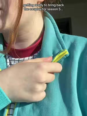A person is shown from the chest up, angled slightly from their left side. They are wearing a teal zip-up jacket with yellow trim around the zipper; under the jacket, they're wearing a red top and a shirt that has a black and white pattern. The person’s face is not fully visible; only part of their cheek and hair are seen. The person's hand is near the zipper, as if they are in the process of zipping it up. The background is blurred and indoors.