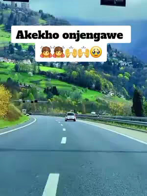 A forward-facing shot taken from inside a vehicle as it travels down a highway. The road curves gently to the left, with a white car driving in the distance. Lush green hills cover the background, and the sky is a muted blue. A white text box with black font is superimposed across the top, and a series of emojis represent various emotions.