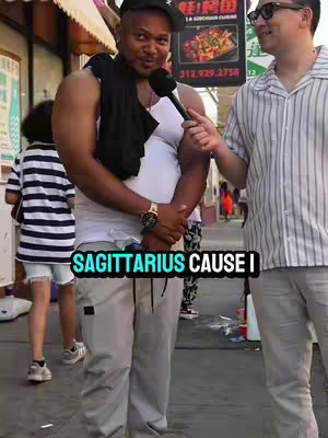 The image shows a man being interviewed on the street. He is wearing a white tank top, grey pants, and a black cap and holding a microphone. The background shows a street scene with a sign.