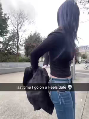 A woman with dark hair, viewed from the back, is standing outdoors. She is wearing a black long-sleeved shirt and blue jeans. She is carrying a black bag. The background shows a pathway with trees and buildings in the distance. Text overlayed at the bottom reads 'last time I go on a Feels date' with a skull emoji.
