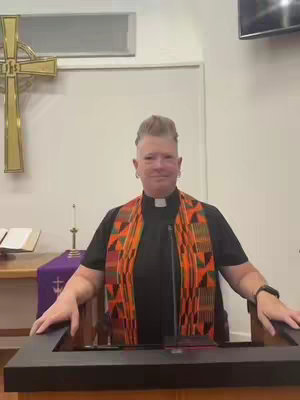 A person, likely a preacher, stands at a podium in a church setting. They are wearing a clerical collar and a patterned scarf, and their hands are resting on the podium. The background includes a wooden cross, a window, and a purple cloth.